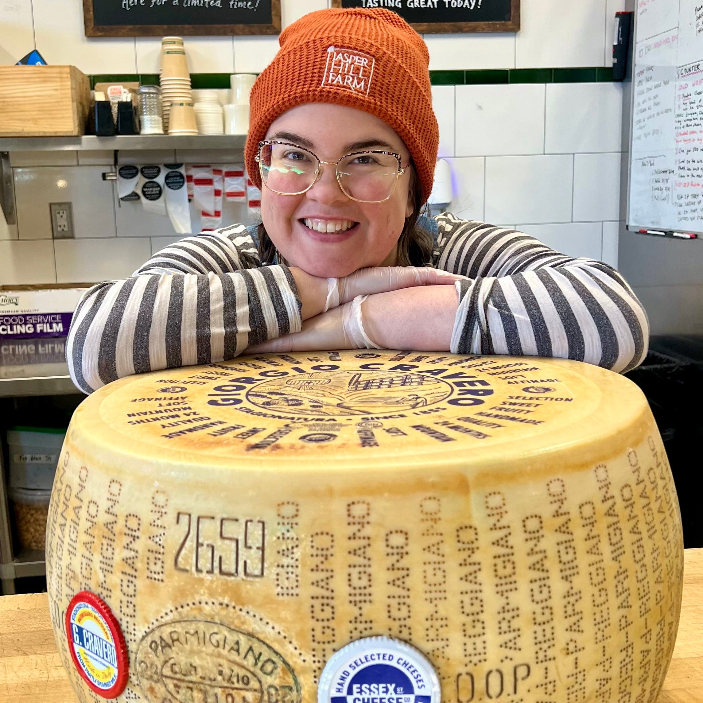 Person wearing an orange beanie and striped sweater sitting on a large wheel of Parmigiano Reggiano cheese.