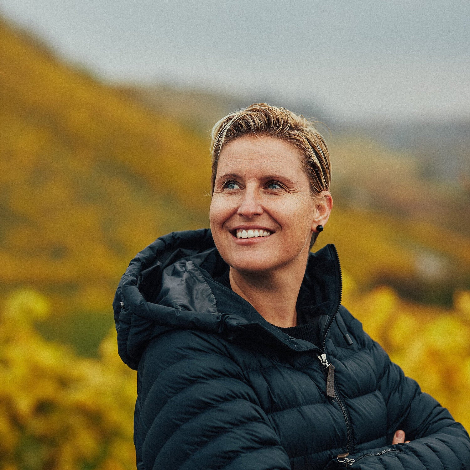 Person wearing a black jacket standing in a scenic outdoor setting with mountains and yellow flowers.