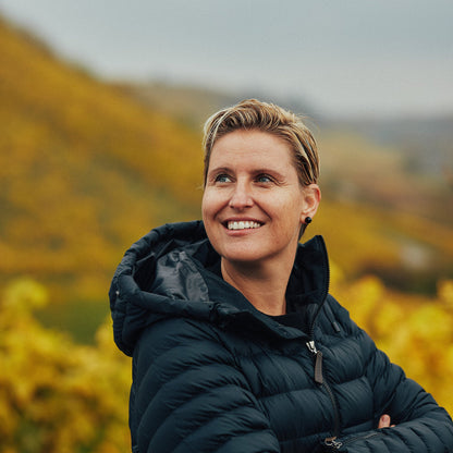 Person wearing a black jacket standing in a scenic outdoor setting with mountains and yellow flowers.