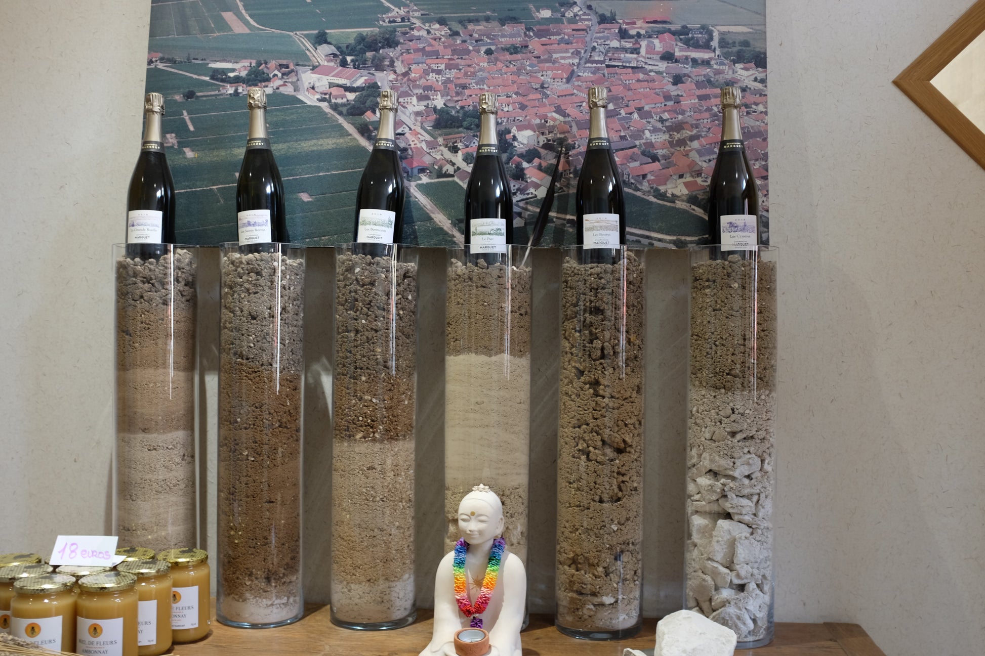 Six tall clear glass cylinders filled with different dried plant materials or grains, arranged on a table in front of a large landscape print. Small jars with gold lids are visible on the left, and a small white figurine stands in front of the cylinders.