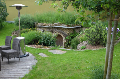A small stone hobbit-style entrance with a grass-covered roof sits in a landscaped garden. A wooden deck with two gray wicker patio chairs and a tall silver patio heater is on the left, with green lawn, shrubs, and trees surrounding the scene.