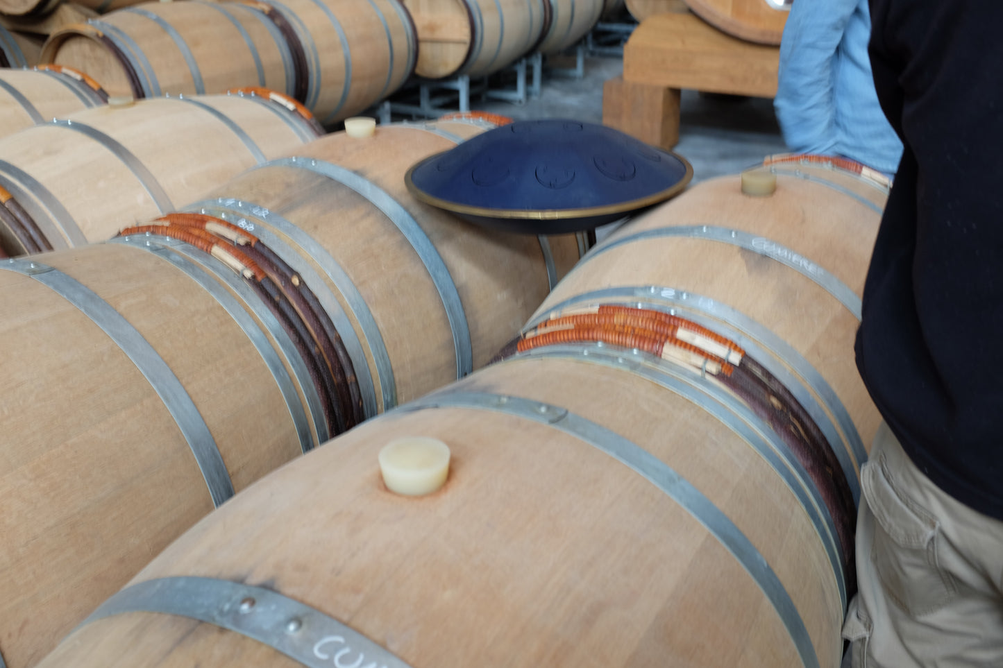 A close-up view of wooden wine barrels with metal bands, with a blue instrument resting on top. A person in a dark navy shirt and khaki pants is partially visible on the right side, standing in a barrel room.