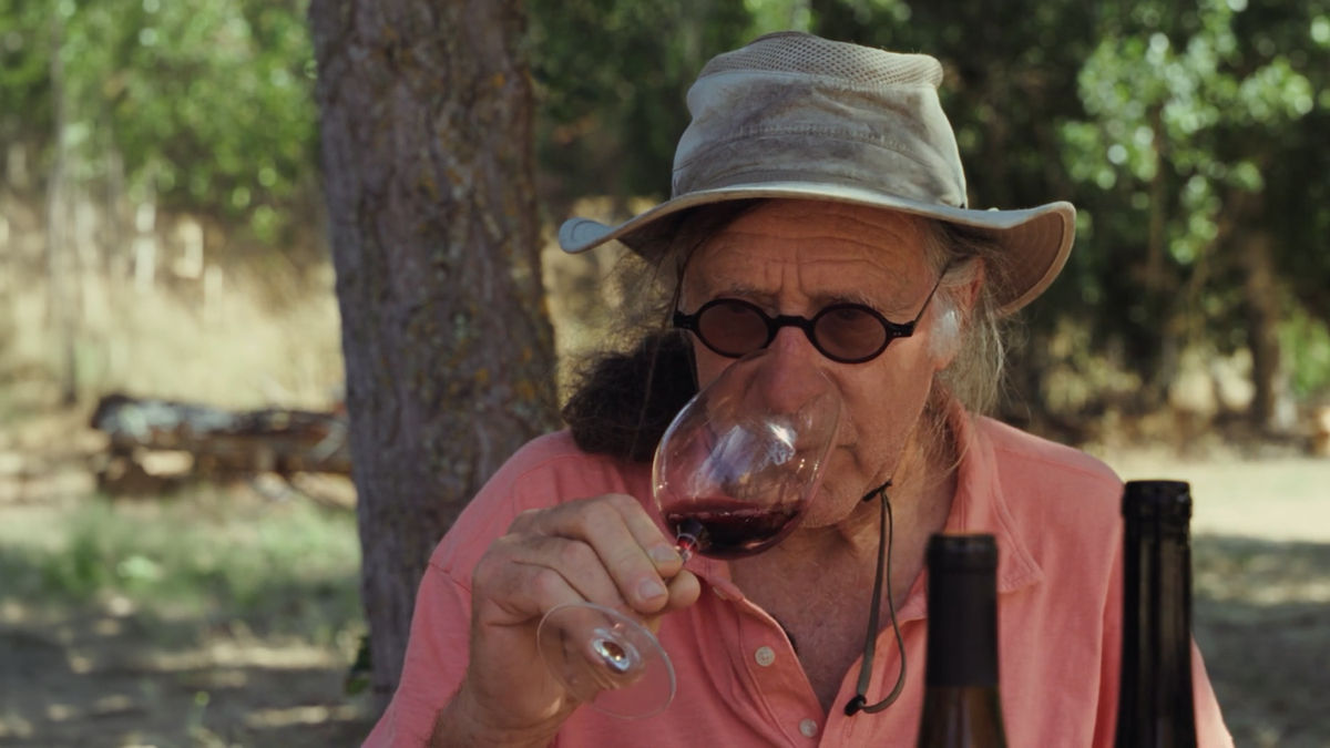 Winemaker Randall Graham samples a glass of red wine. He wears a tan hat and a small pair of sunglasses. 