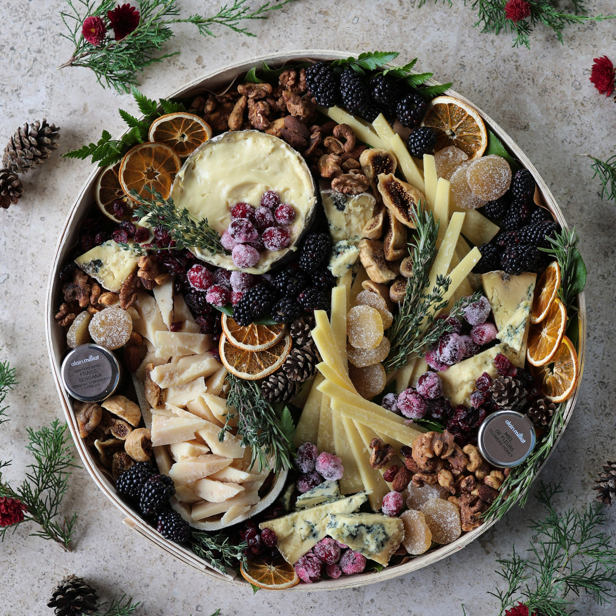 A top-down view of a round charcuterie board filled with cheeses, nuts, dried orange slices, blackberries, and sprigs of rosemary, arranged on a light stone surface with pine branches around the edges.