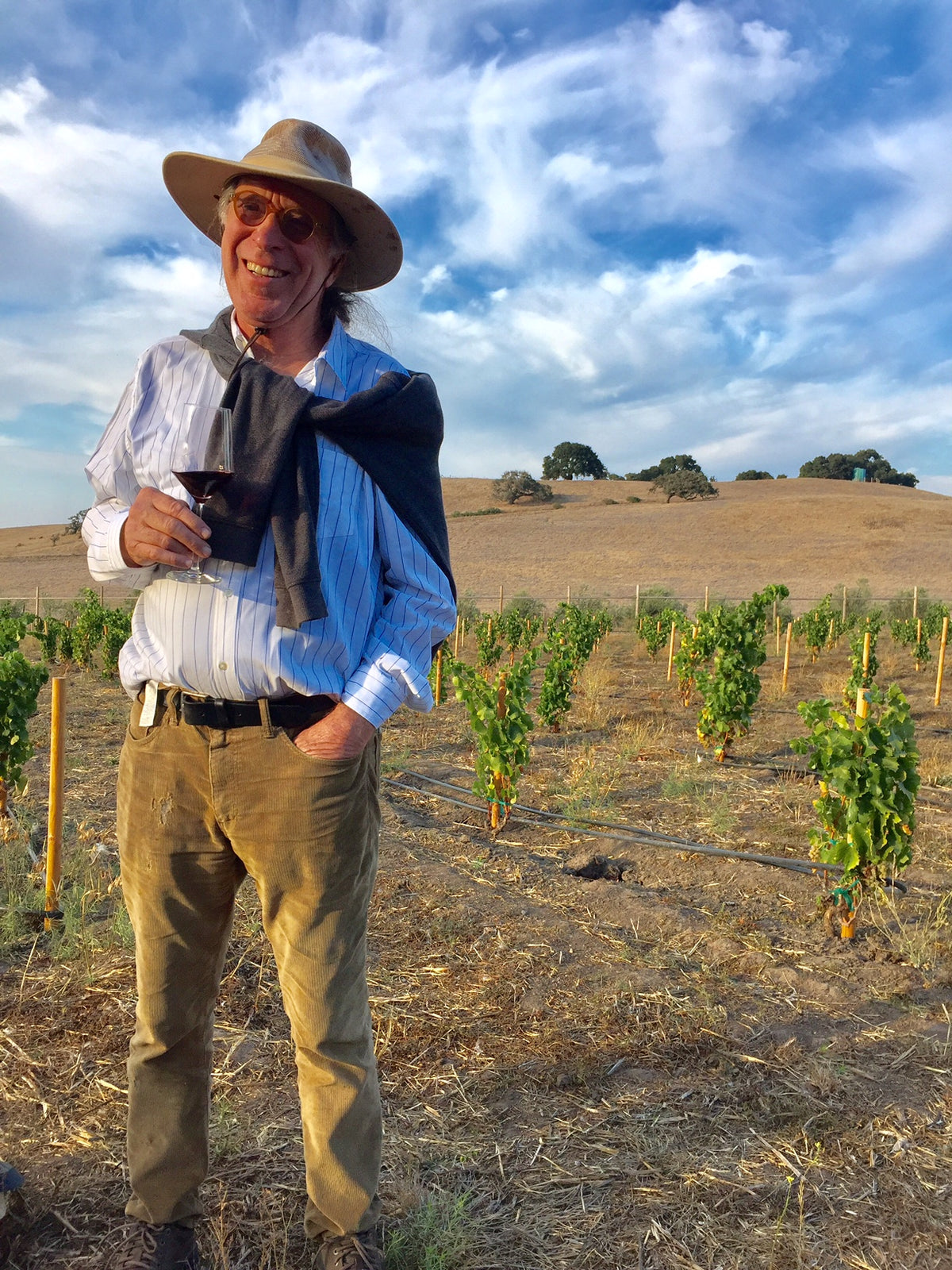 Winemaker Randall Graham stands in front of waist-high grape vines, holding a glass of red wine. He wears a striped button-up shirt and tan pants and hat, with a black sweater loosely tied around his shoulders.
