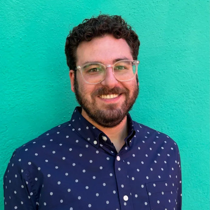 A man in glasses with a brown beard and curly hair stands in front of a bright turquoise wall