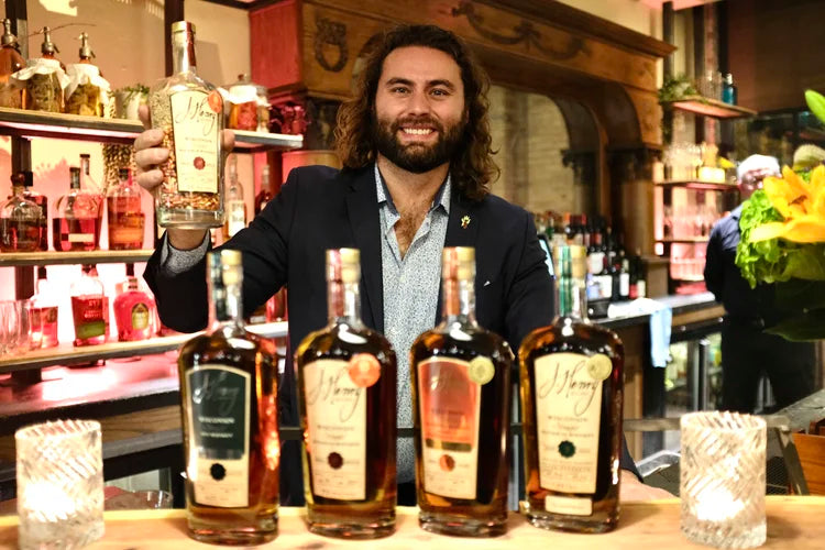 A smiling man with long dark hair stands behind a bar counter holding a bottle in one hand, with four bottles of whiskey lined up in front of him. 