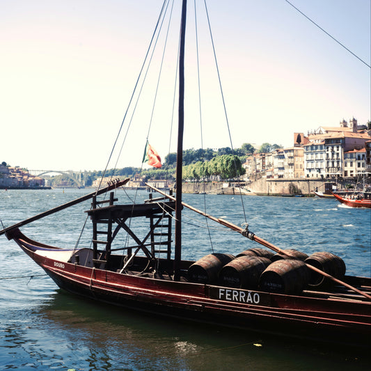 A traditional Portuguese ruyta boat with barrels on deck, labeled 'FERRAO', floating on the Douro River with Porto's cityscape in the background.